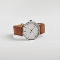 Alcott luxury watch with polished silver round dial, minimalist black index markers, and cognac brown genuine leather strap with precision stitching, displayed at three-quarter angle on neutral background