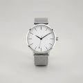 Silver watch with round white dial and black hour markers, displayed flat against neutral background showing full face view with minimalist design